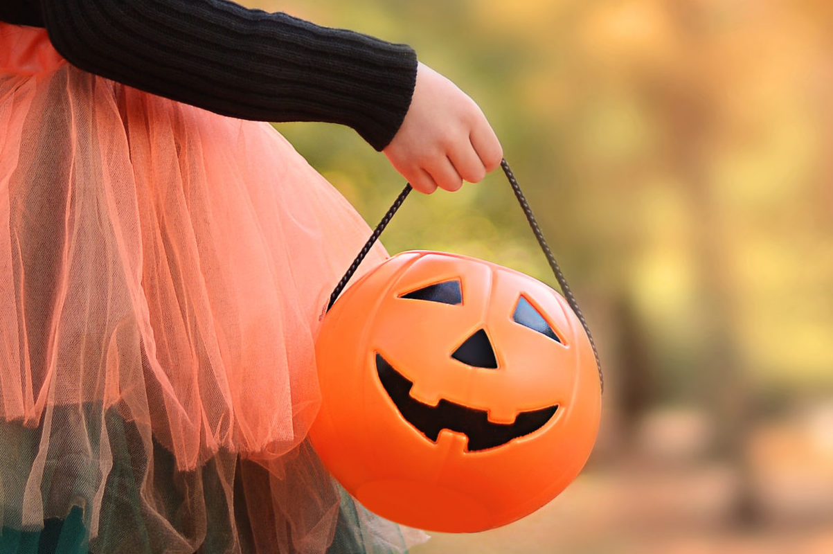 Little girl in witch costume with a balloon and a pumpkin bucket playing in autumn park. Child having fun at Halloween trick or treat. Kids trick or treating. Banner. Copy space
